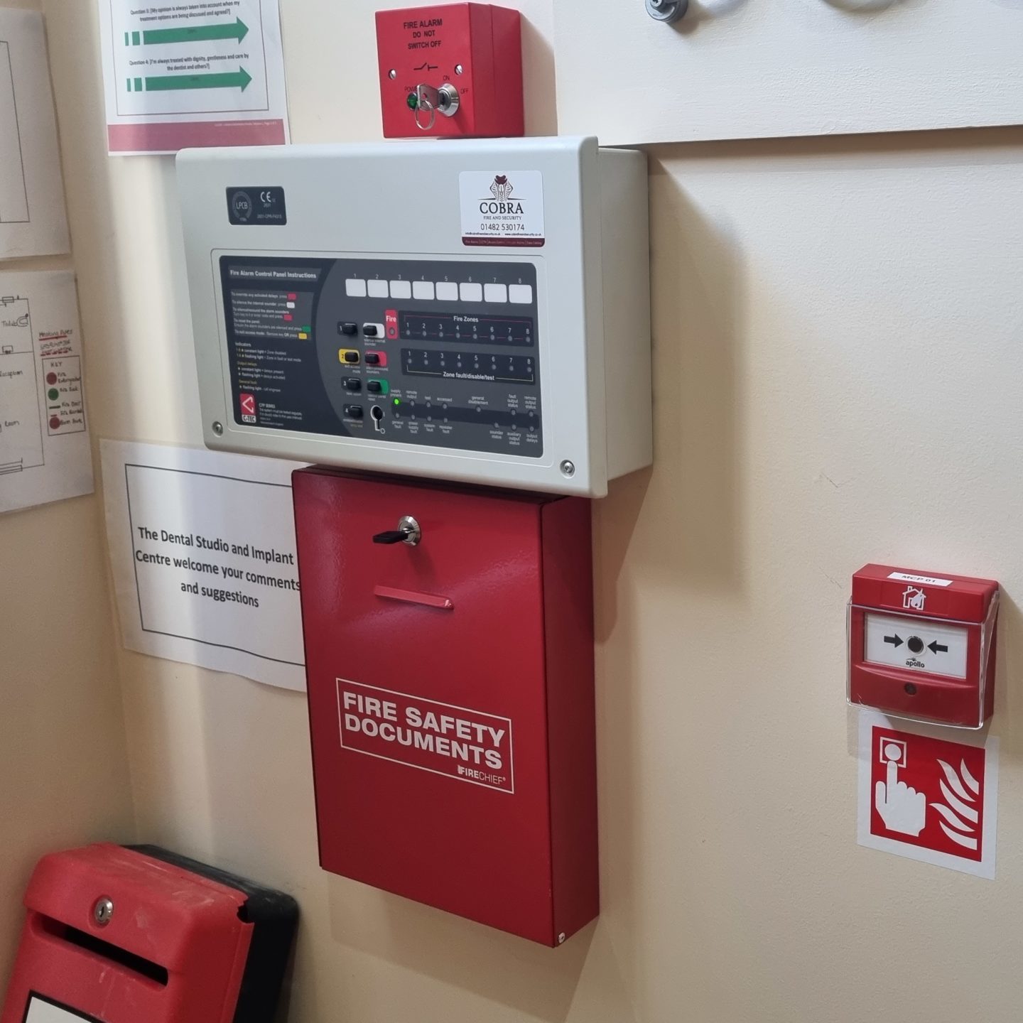 A fire Alarm mounted on a wall next to a fire alarm call point and documents box for fire alarm documents.