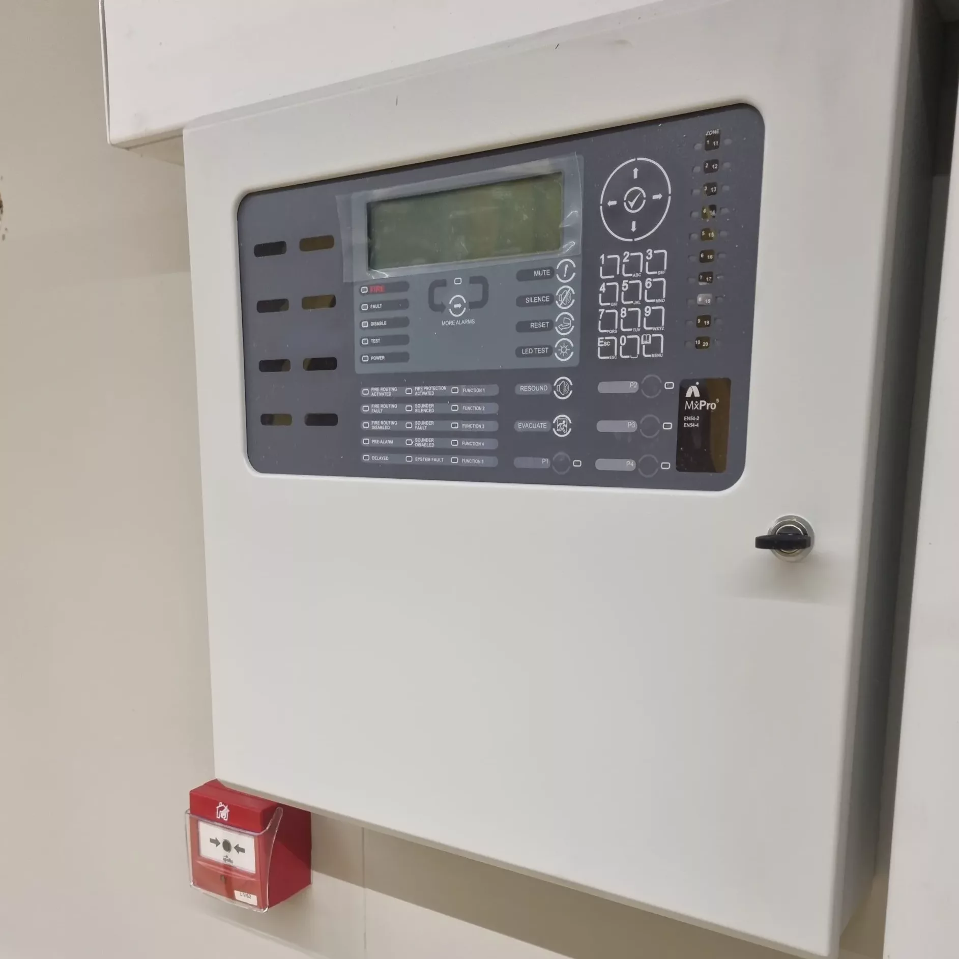 Wall-mounted fire alarm control panel with a red manual call point installed directly below it.