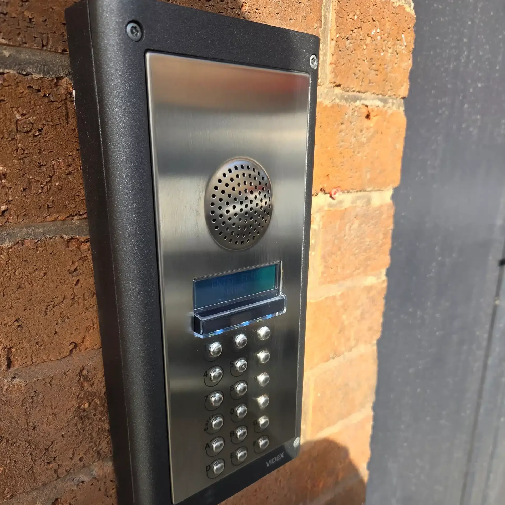 Videx audio intercom entry panel with keypad and digital display, installed on a brick wall at the entrance to a block of flats